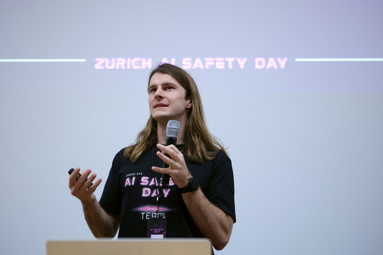 A speaker with a microphone in front of a Zurich AI Safety Day title slide